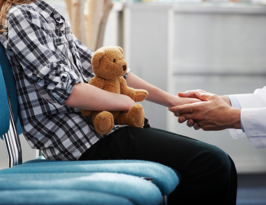 Patient sitting in lobby holding teddy bear being comforted by doctor